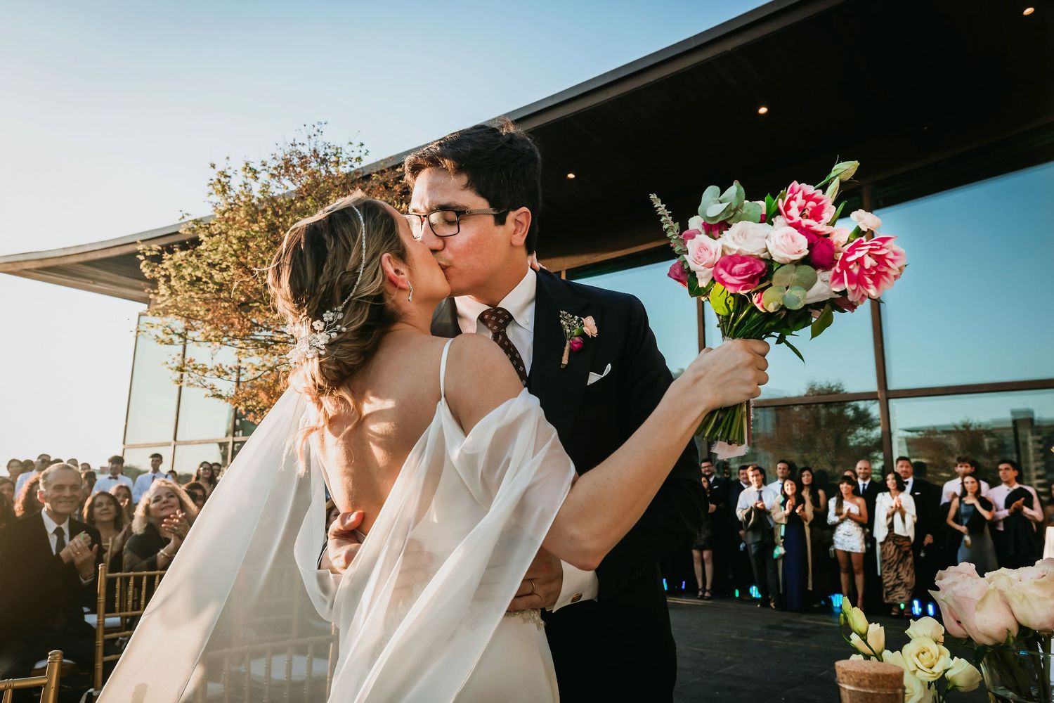 Newlyweds share first kiss holding vibrant pink bouquet at outdoor wedding ceremony.