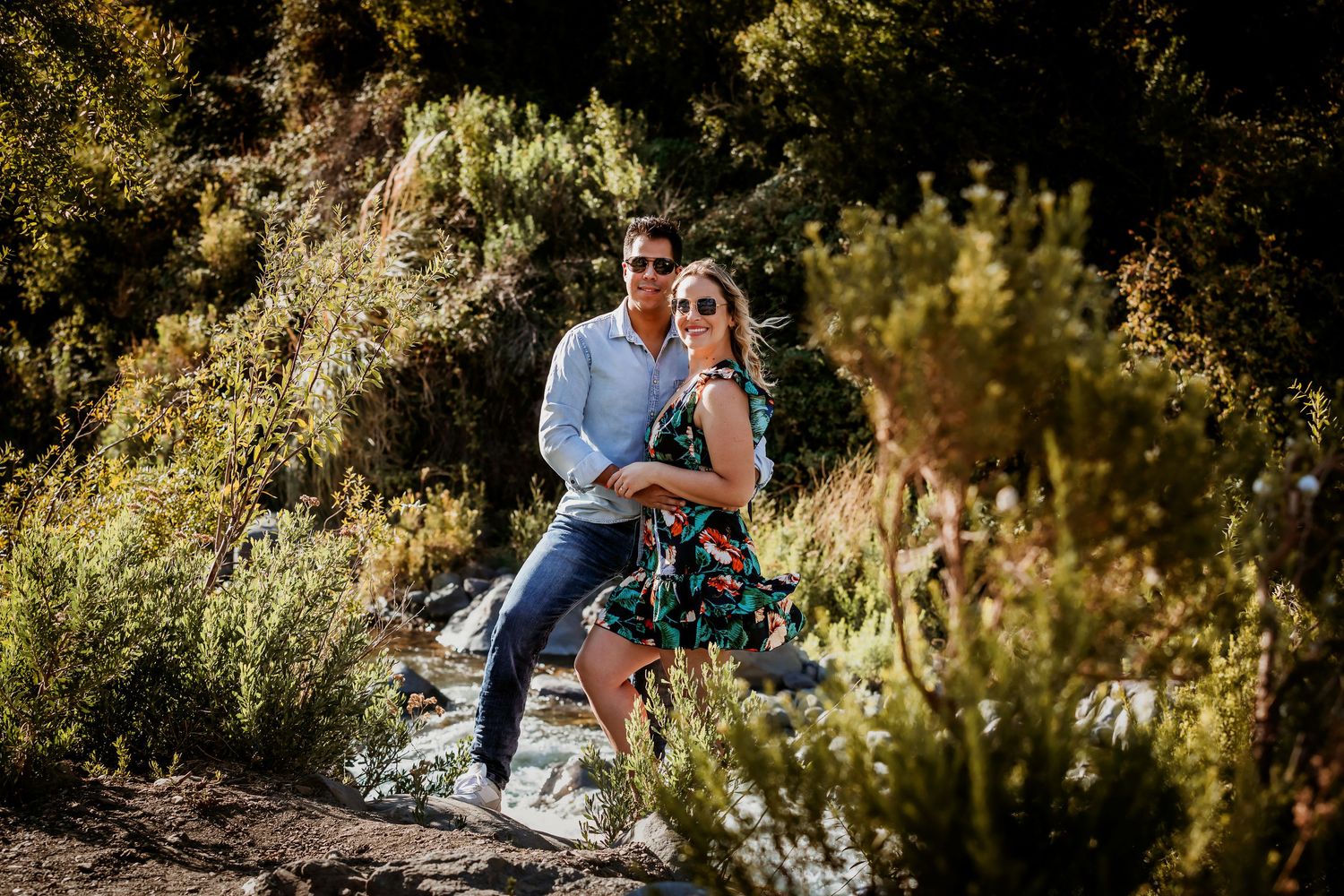 Couple posing together in a lush outdoor setting, woman in floral dress and man in blue shirt, surrounded by green shrubs.
