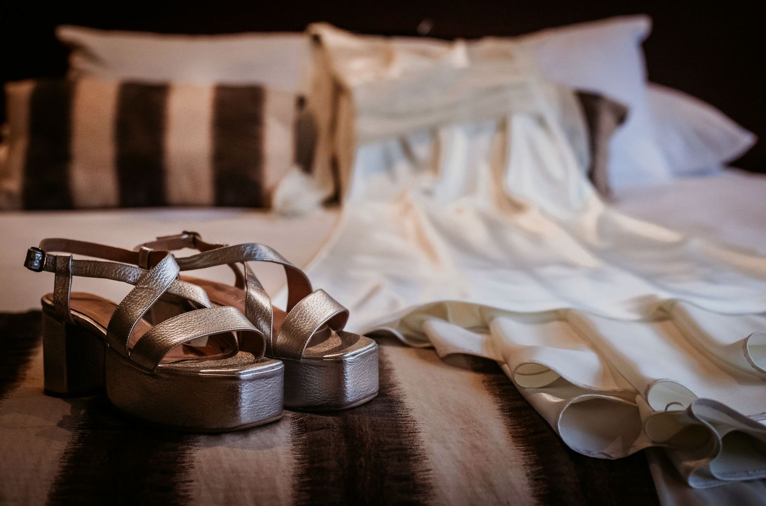 Elegant silver platform sandals displayed on a wooden surface with a white wedding dress draped on a bed in the background.
