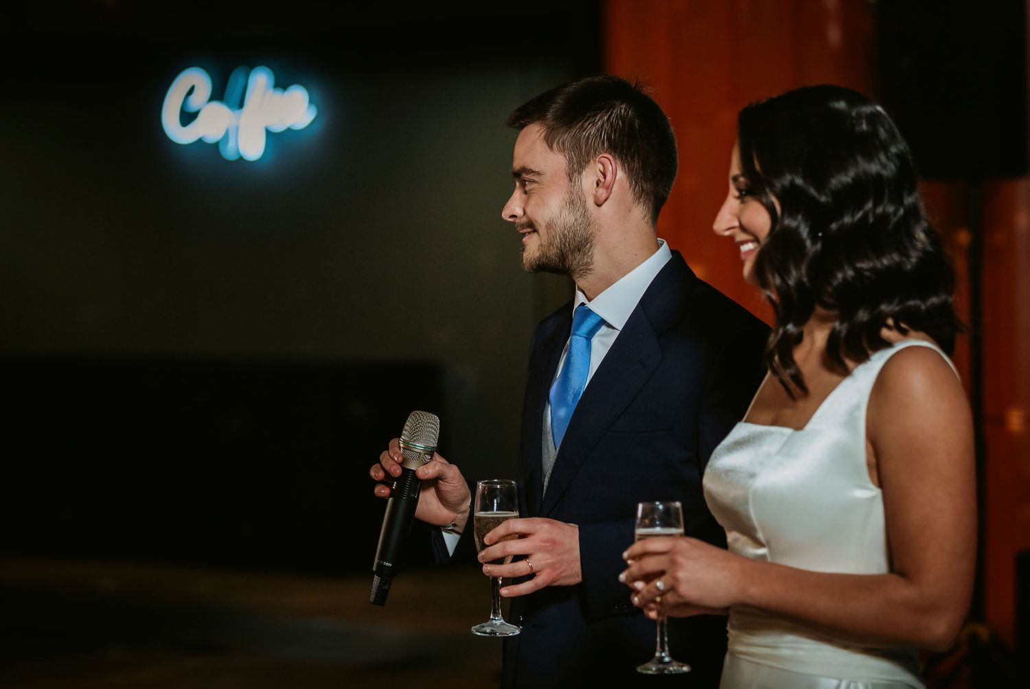A smiling bride and groom holding champagne glasses during their wedding reception with a glowing neon sign in the background.