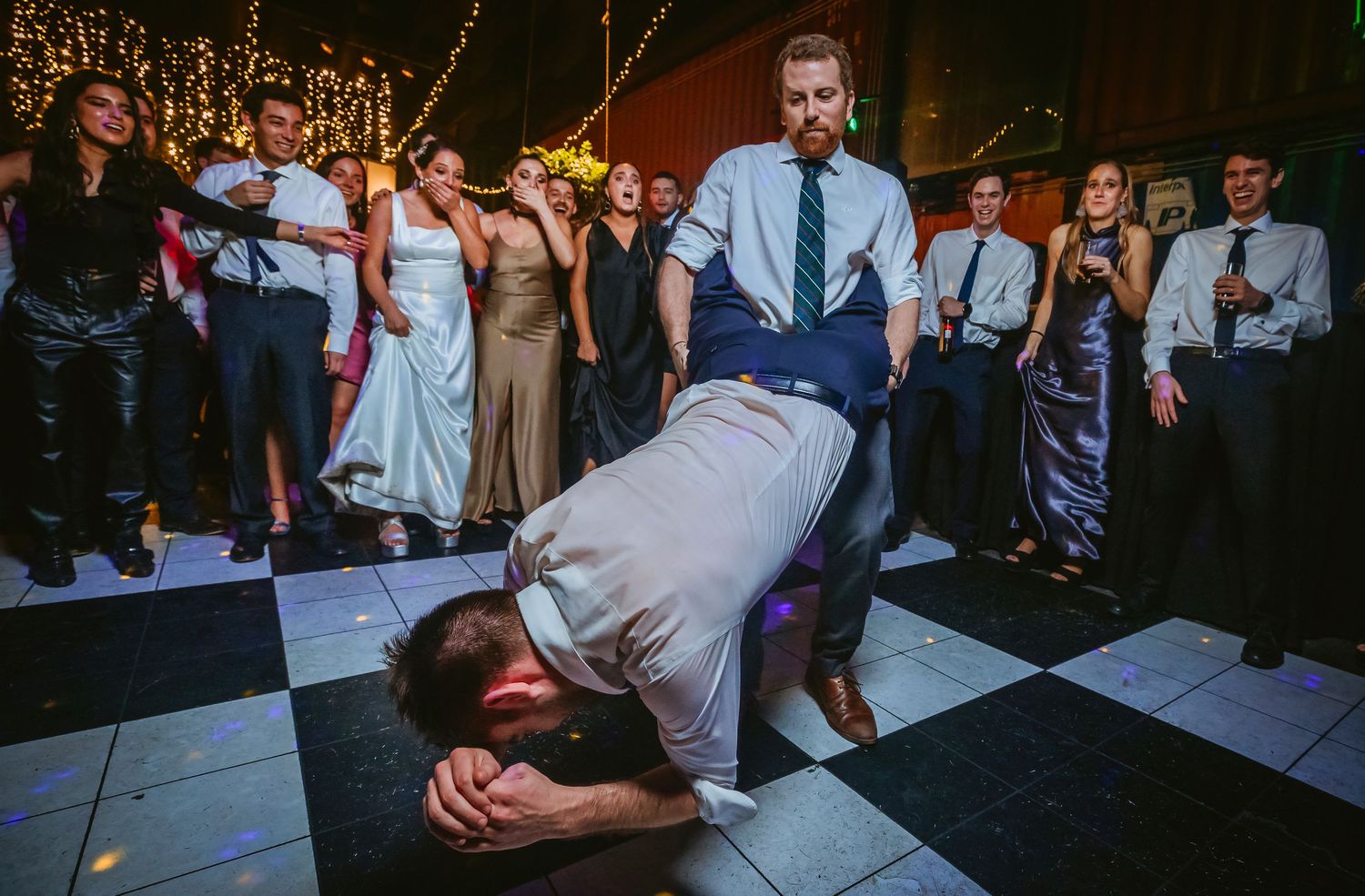 Energetic wedding guest performs dance move on checkered floor while crowd watches at reception.