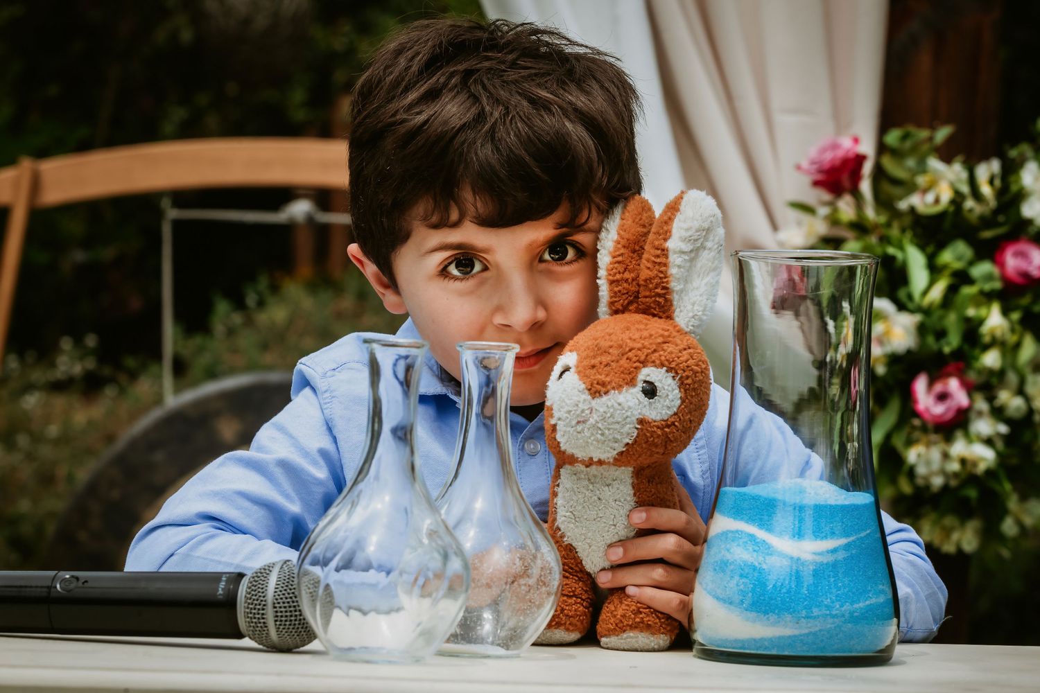 Young boy holding a stuffed bunny toy at a table with a microphone and glass vases at an outdoor event.