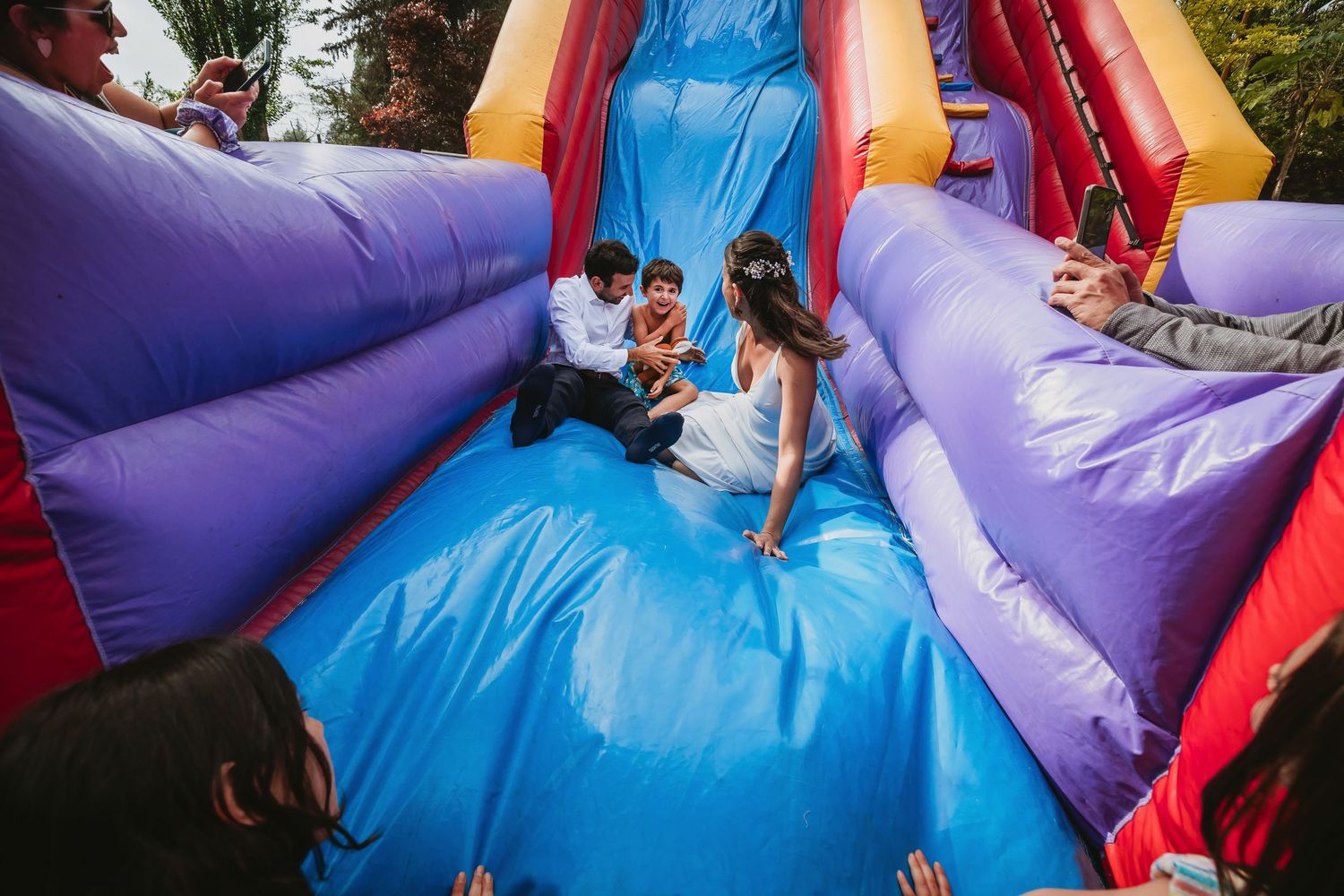 Bride and groom sliding down a colorful inflatable bounce house slide together at a fun outdoor wedding celebration.