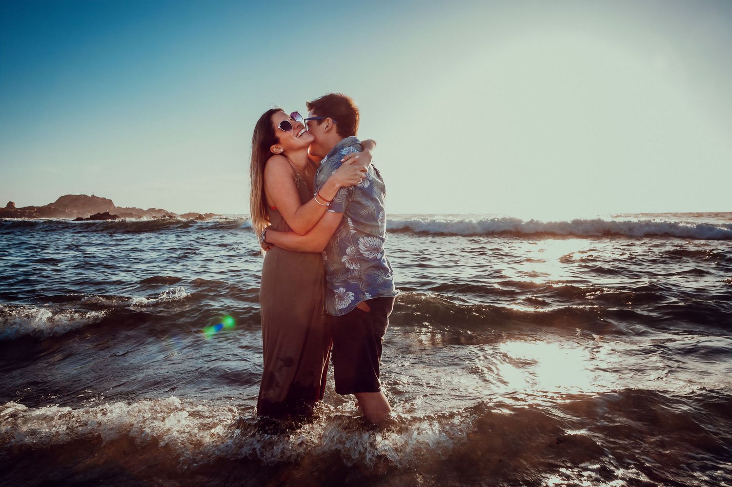 Couple kissing romantically in ocean waves at sunset on a rocky beach.