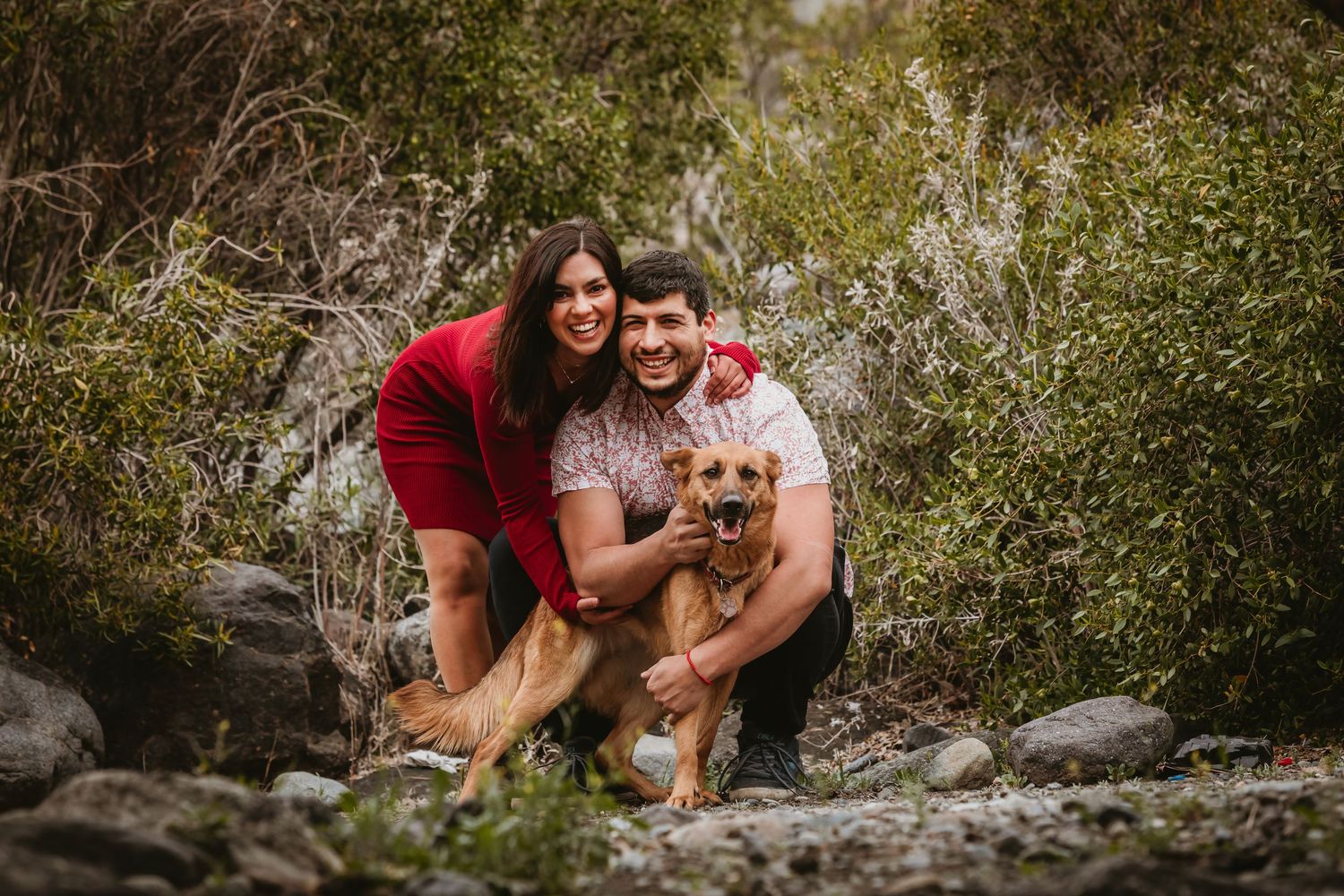 Smiling couple crouching with their small dog among rocks and green shrubs outdoors.