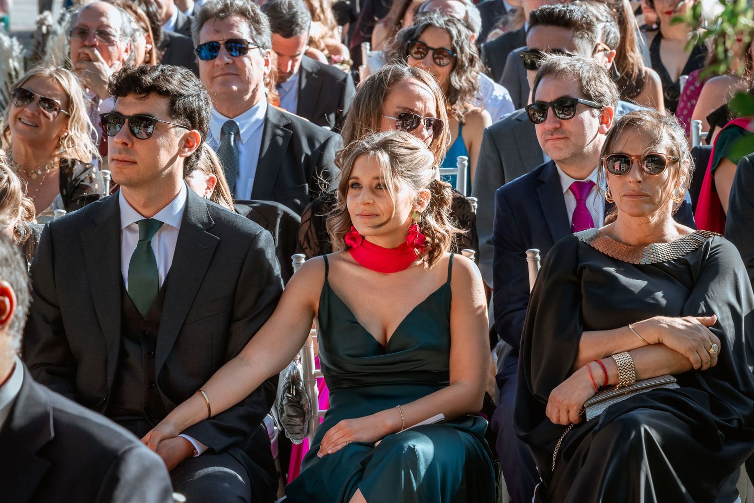 Wedding guests in formal attire seated at sunny outdoor ceremony.