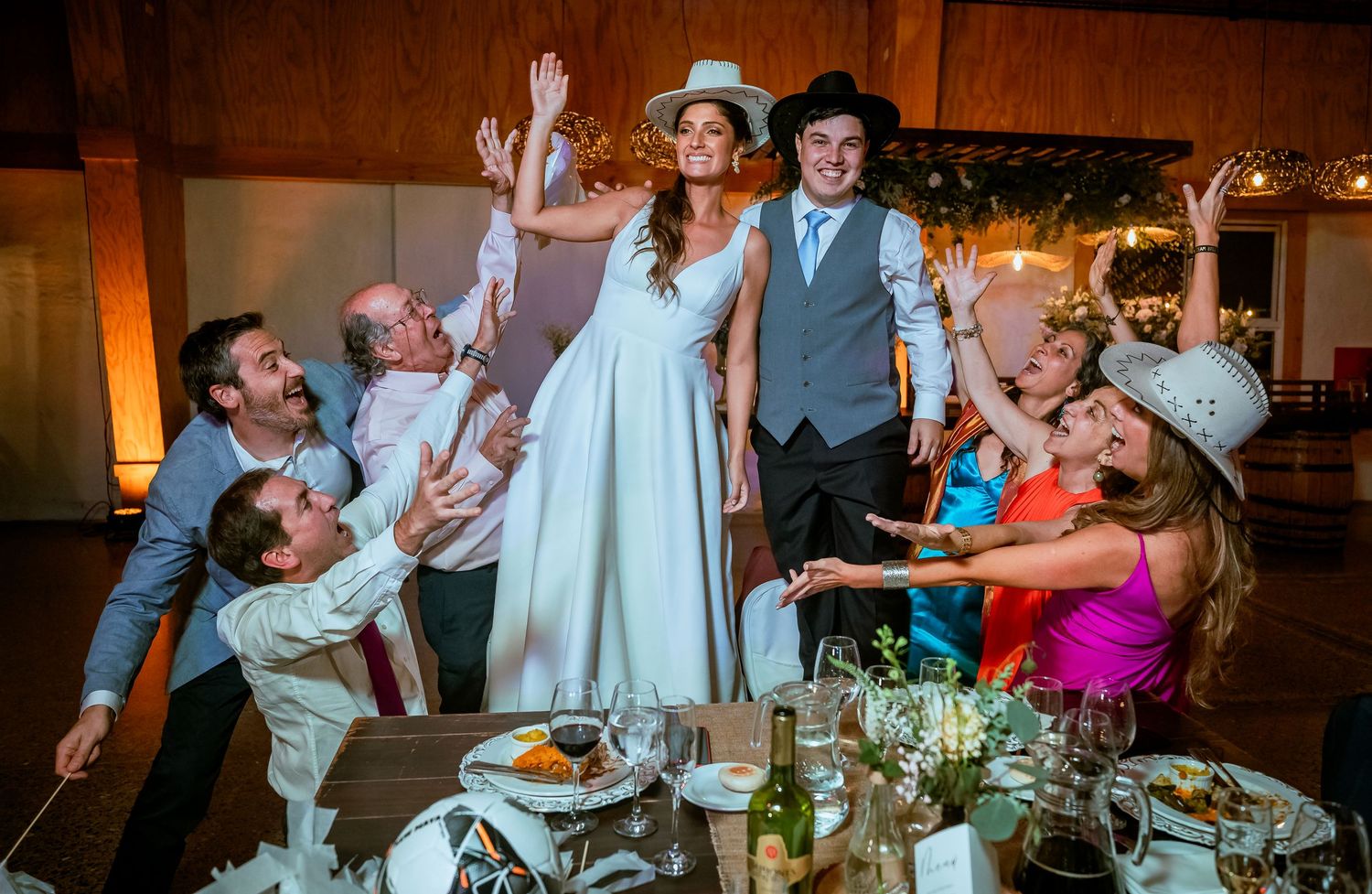 Bride stands on table surrounded by cheering guests at festive wedding celebration.