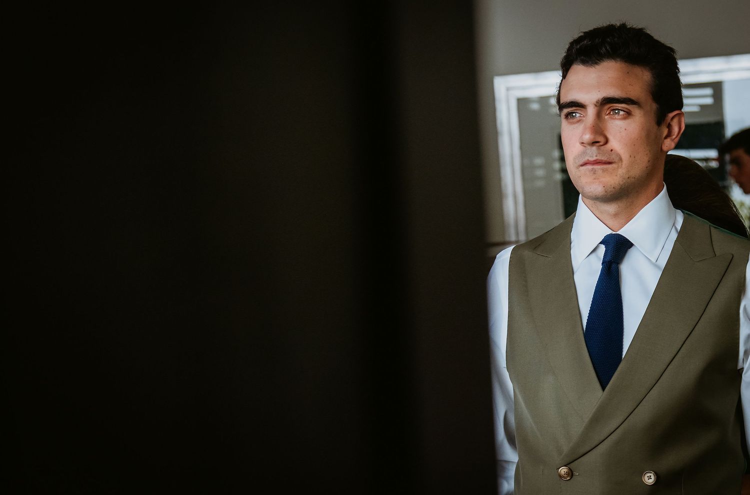 Elegant groom in olive double-breasted suit and navy tie standing in soft light.