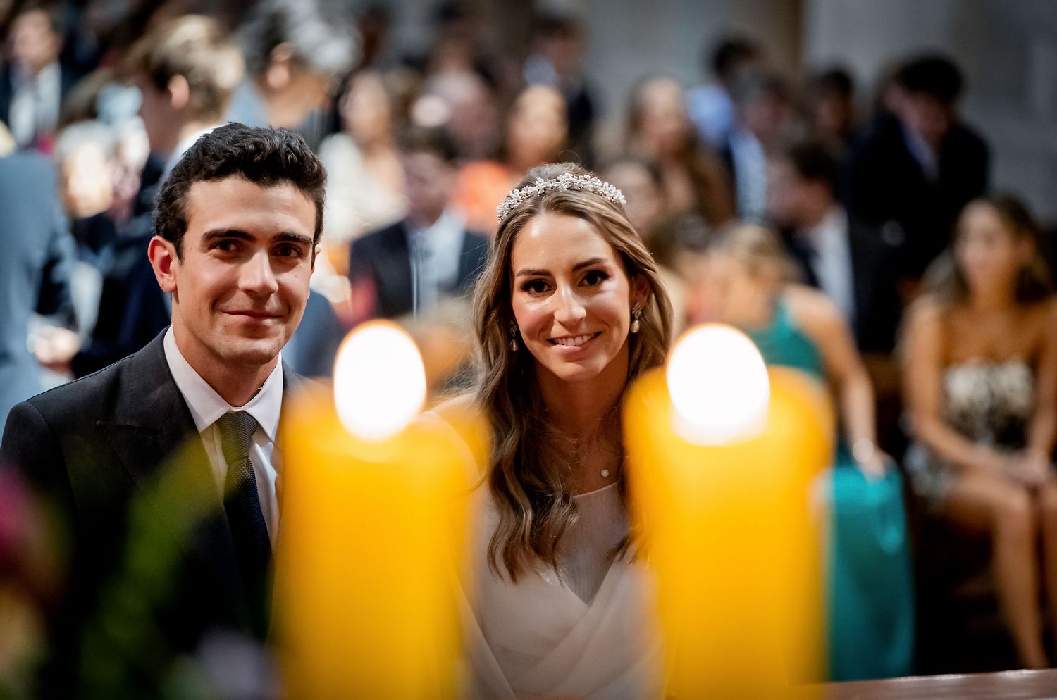 Smiling bride and groom at church altar with large lit candles in foreground and guests seated behind them.
