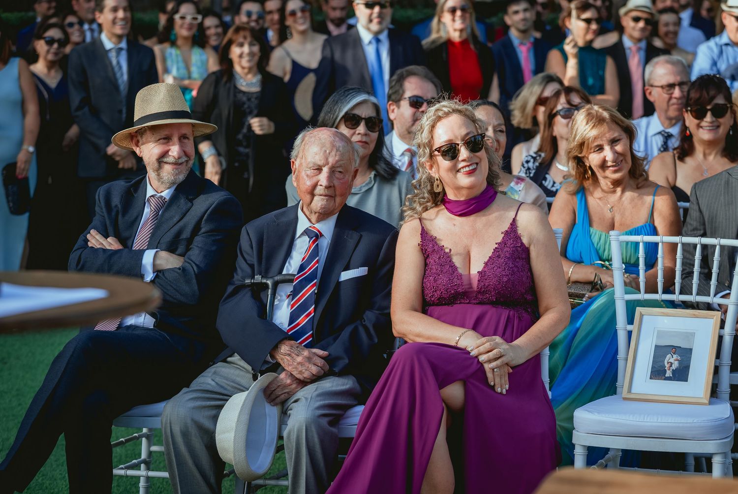 Elegantly dressed wedding guests seated outdoors, including elderly man in suit and woman in purple dress.