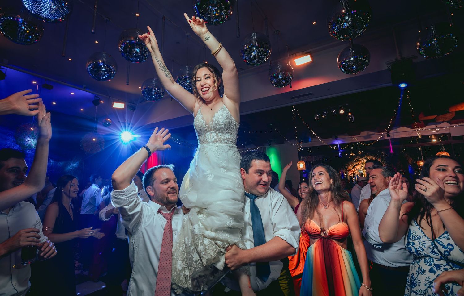 Joyful bride raised by guests on crowded wedding reception dance floor with disco ball lighting.