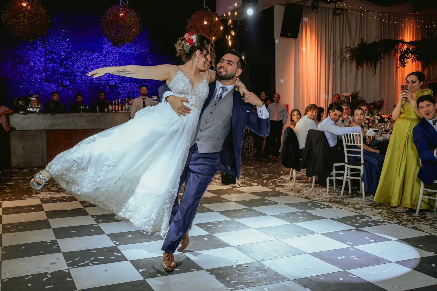 Groom lifts bride in stunning dance move on checkered floor at vibrant wedding reception with blue lighting.