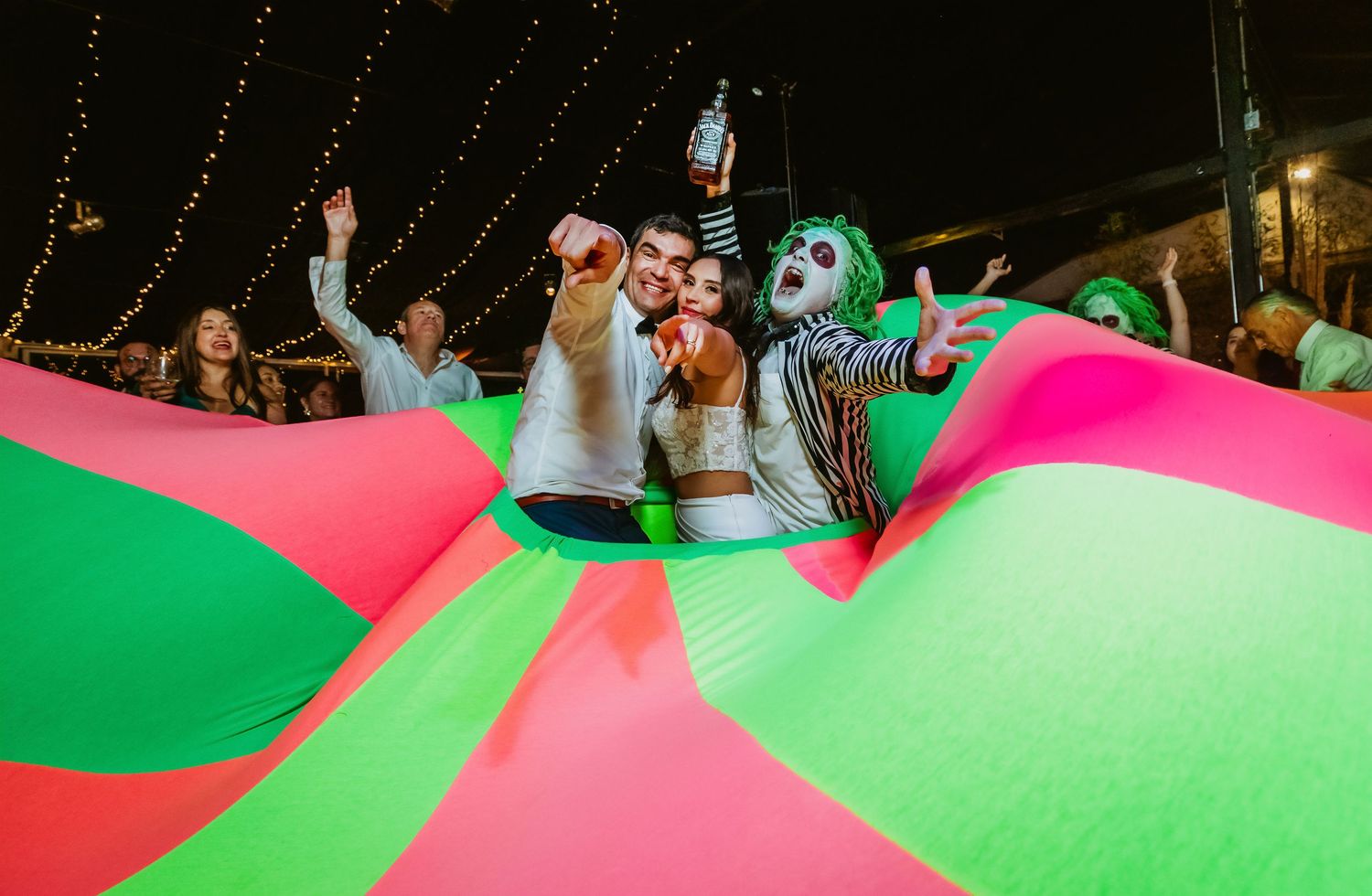 People celebrating at a colorful party with a green and pink parachute, one person dressed as a clown joker at night.