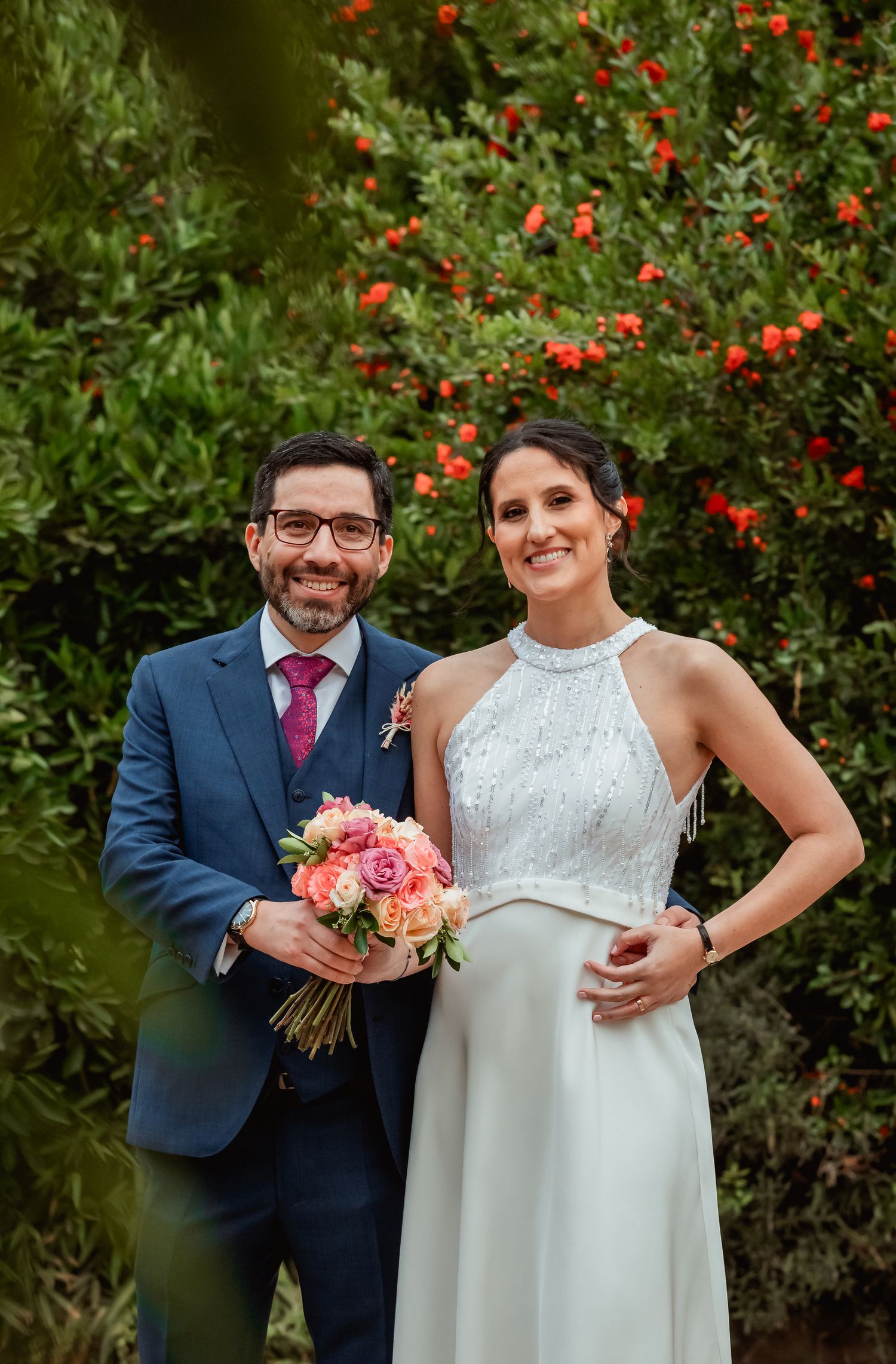 Smiling wedding couple posing in front of lush greenery with red berries, bride holding a pink rose bouquet.