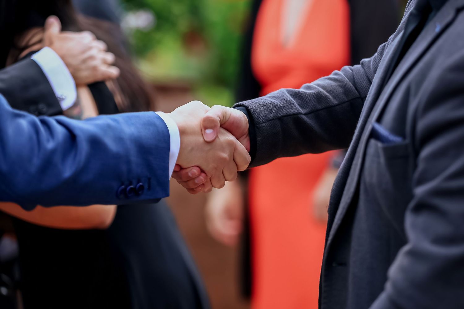 Two men in suits sharing a firm handshake at an outdoor wedding celebration.