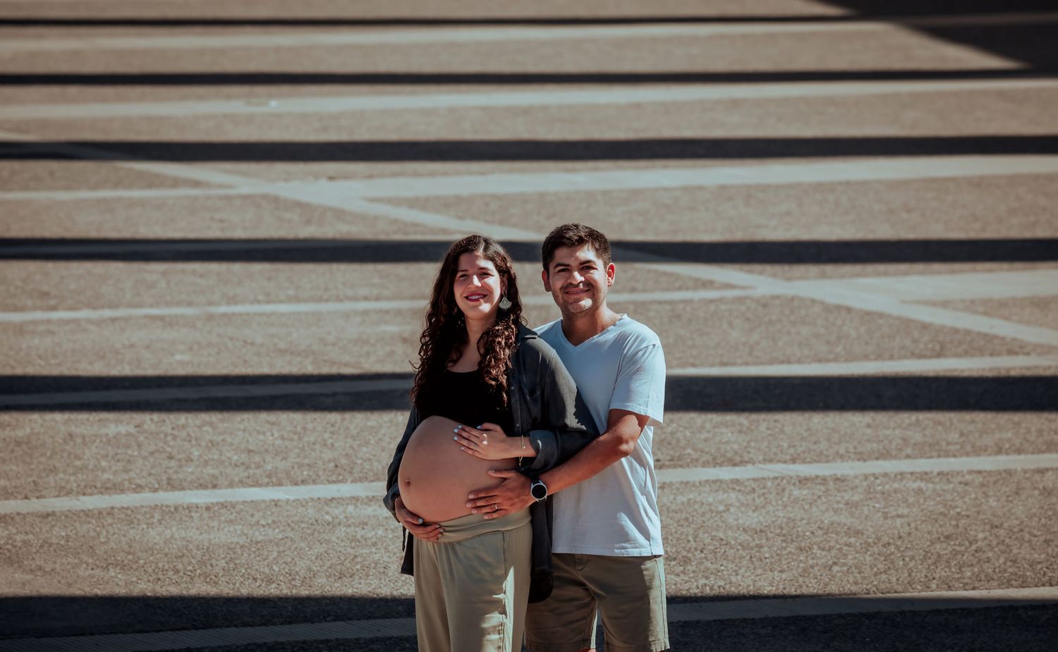 Happy couple embracing on a sunny plaza, man gently holding pregnant woman's bare baby bump.