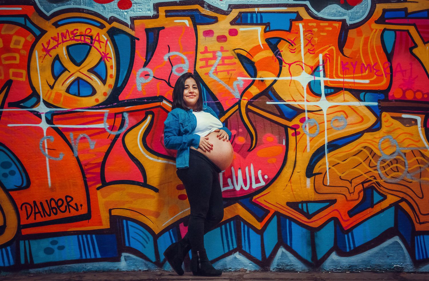 Pregnant woman in denim jacket posing confidently in front of vibrant colorful graffiti wall.