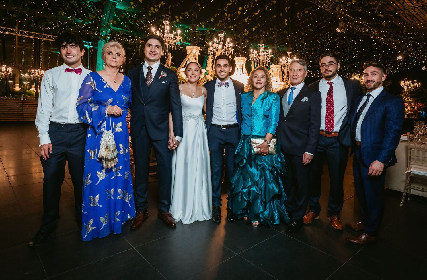 Wedding party group photo of nine guests posing under fairy lights at reception.