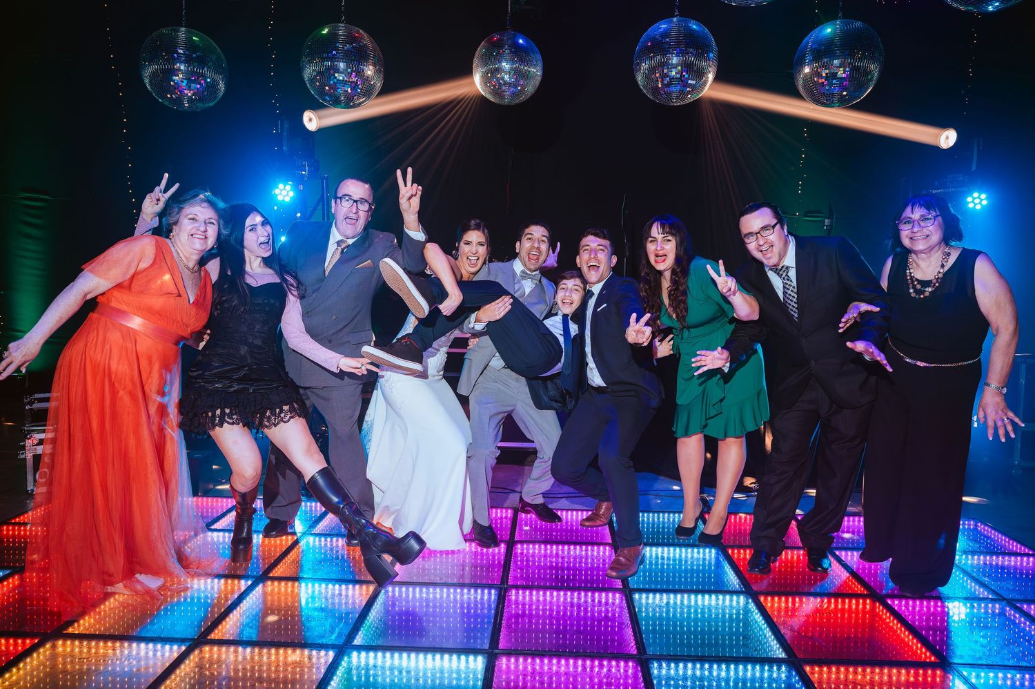 Group of wedding guests posing on colorful LED dance floor under disco balls in a festive venue.