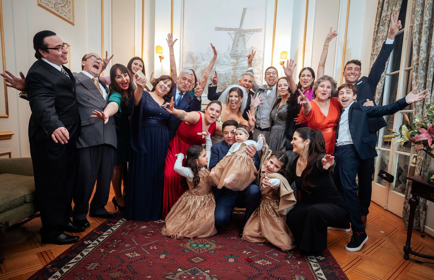 Large joyful wedding party group photo in elegant room with windmill painting, guests celebrating with raised arms.