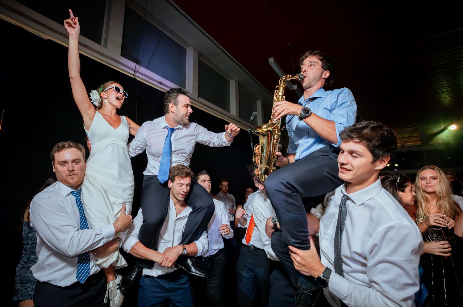 Bride lifted by groomsmen while saxophonist performs at energetic wedding reception dance floor.