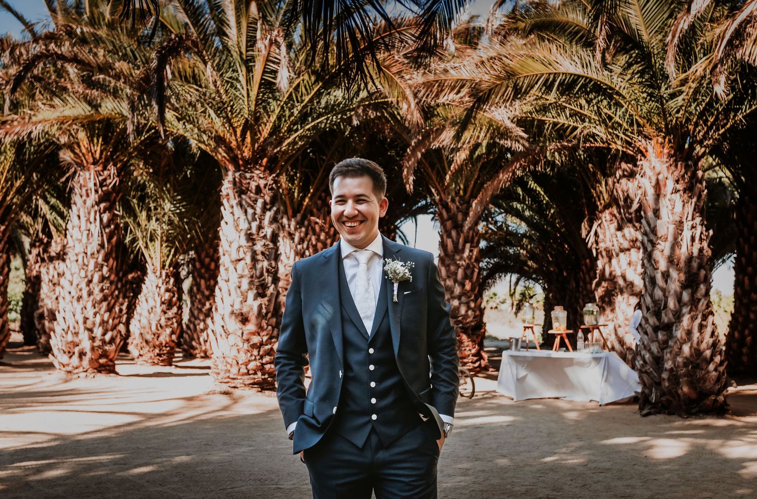 Smiling groom in navy blue suit standing among tall palm trees at outdoor wedding venue.