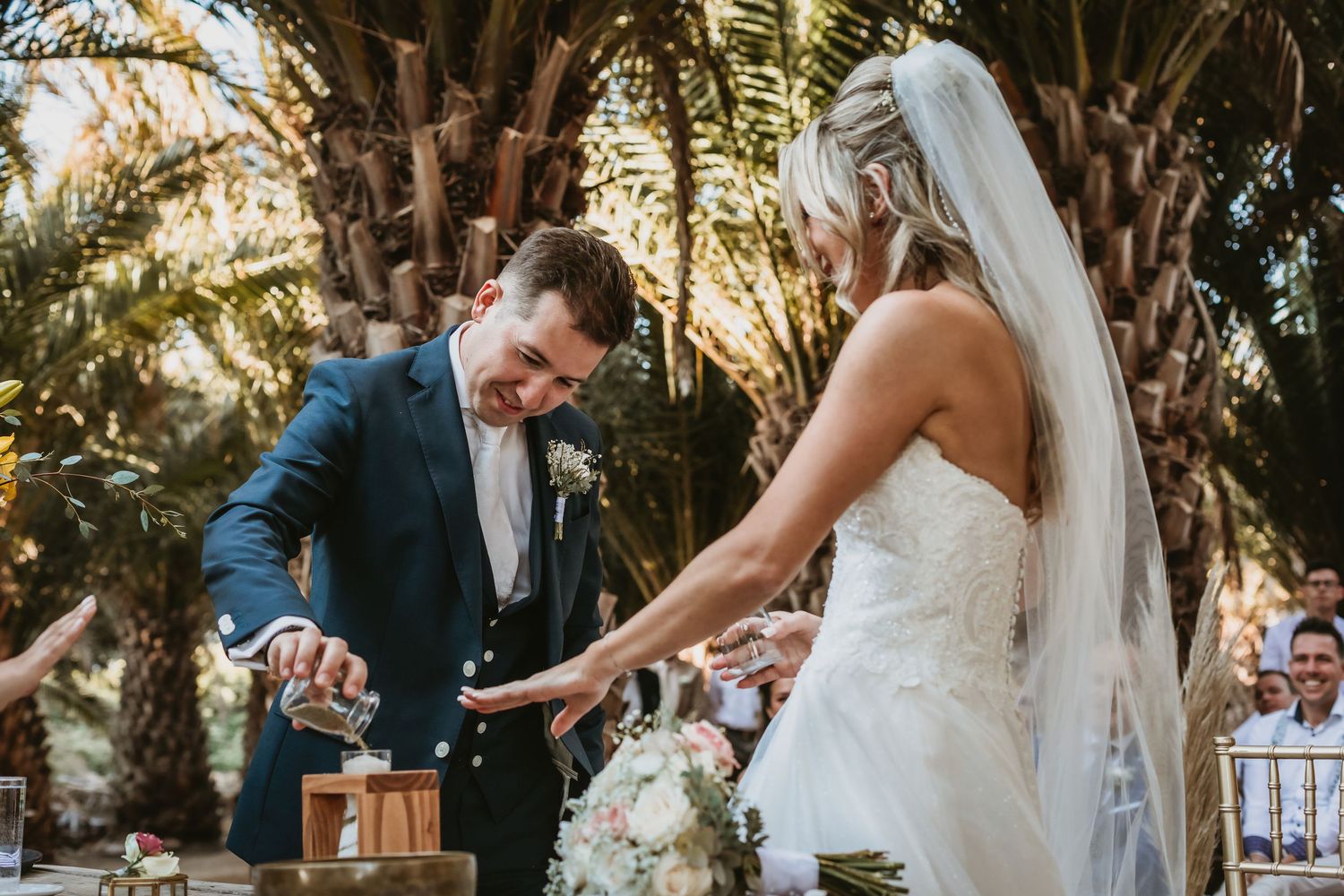 Bride and groom perform a sand unity ceremony at their outdoor wedding surrounded by lush palm trees.