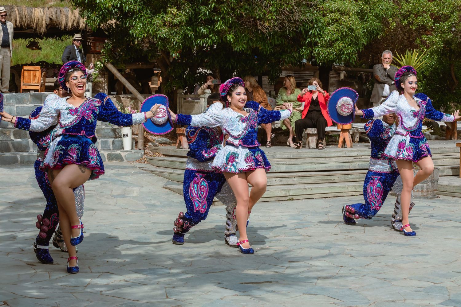 Colorful traditional dancers in vibrant blue and purple costumes performing folk dance on an outdoor stone plaza.