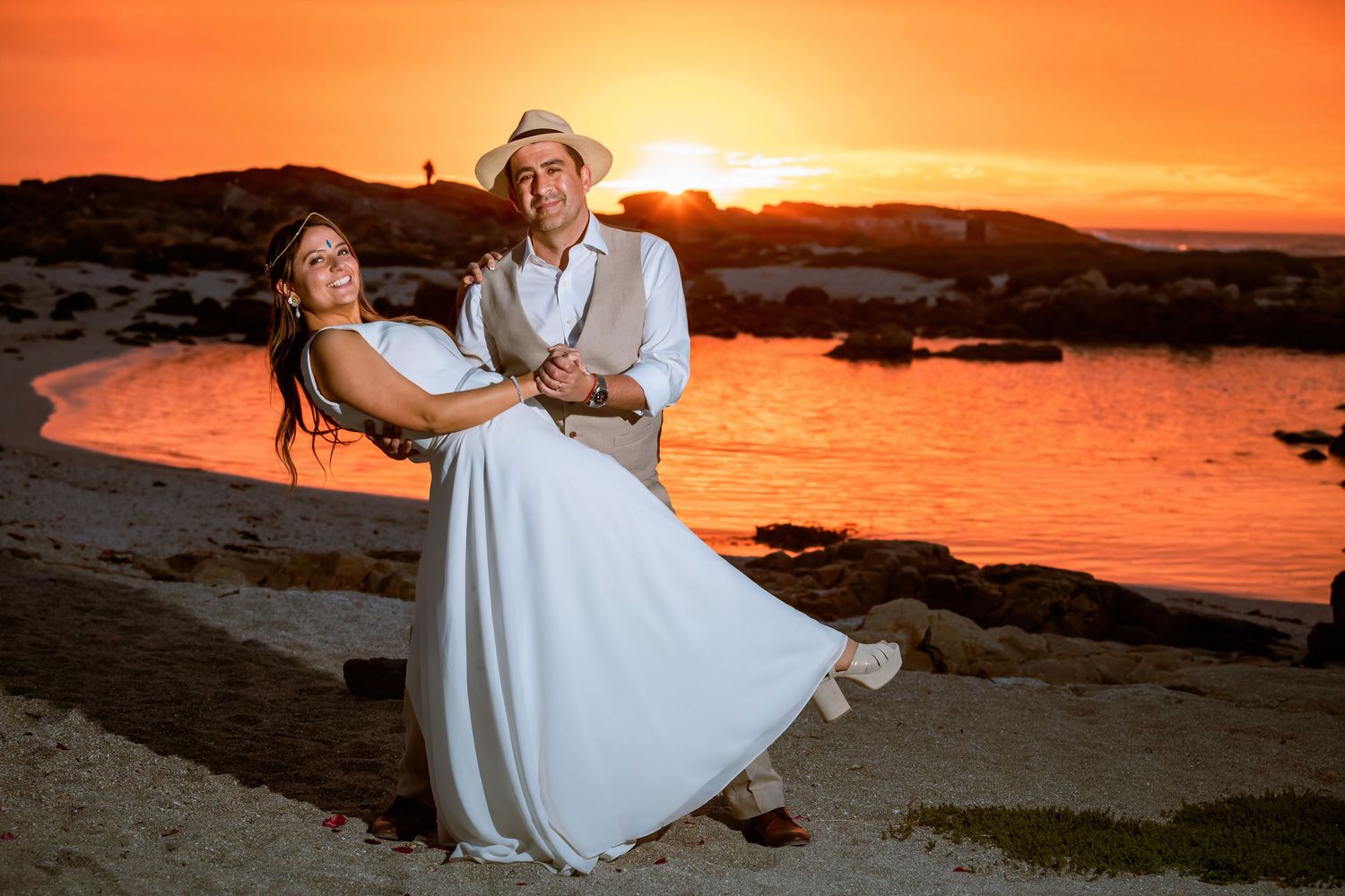 Groom lifting bride in a white dress against a vivid orange sunset on a scenic rocky beach.