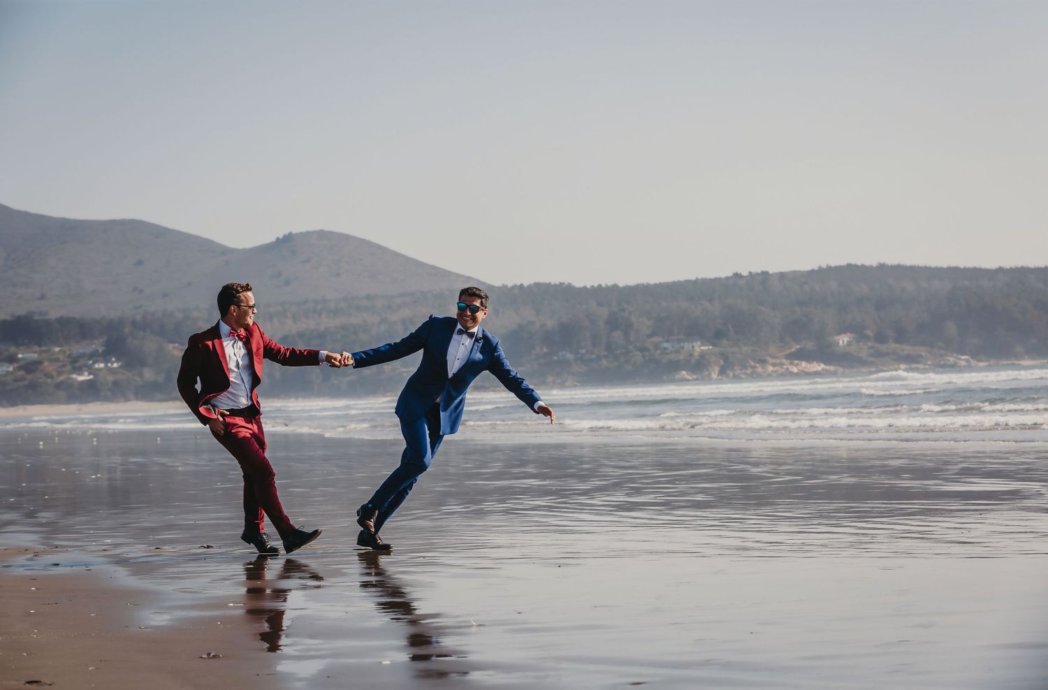 Two grooms in red and blue suits hold hands running on a scenic beach with mountains in the background.