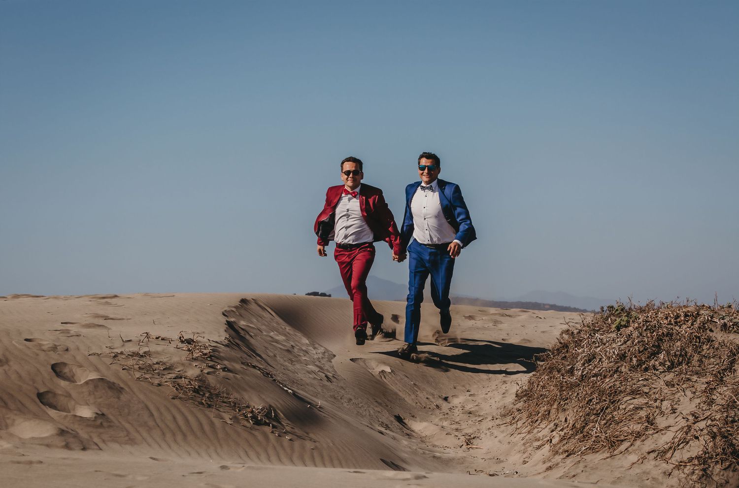 Two grooms in red and blue suits running joyfully across sandy desert dunes on their wedding day.