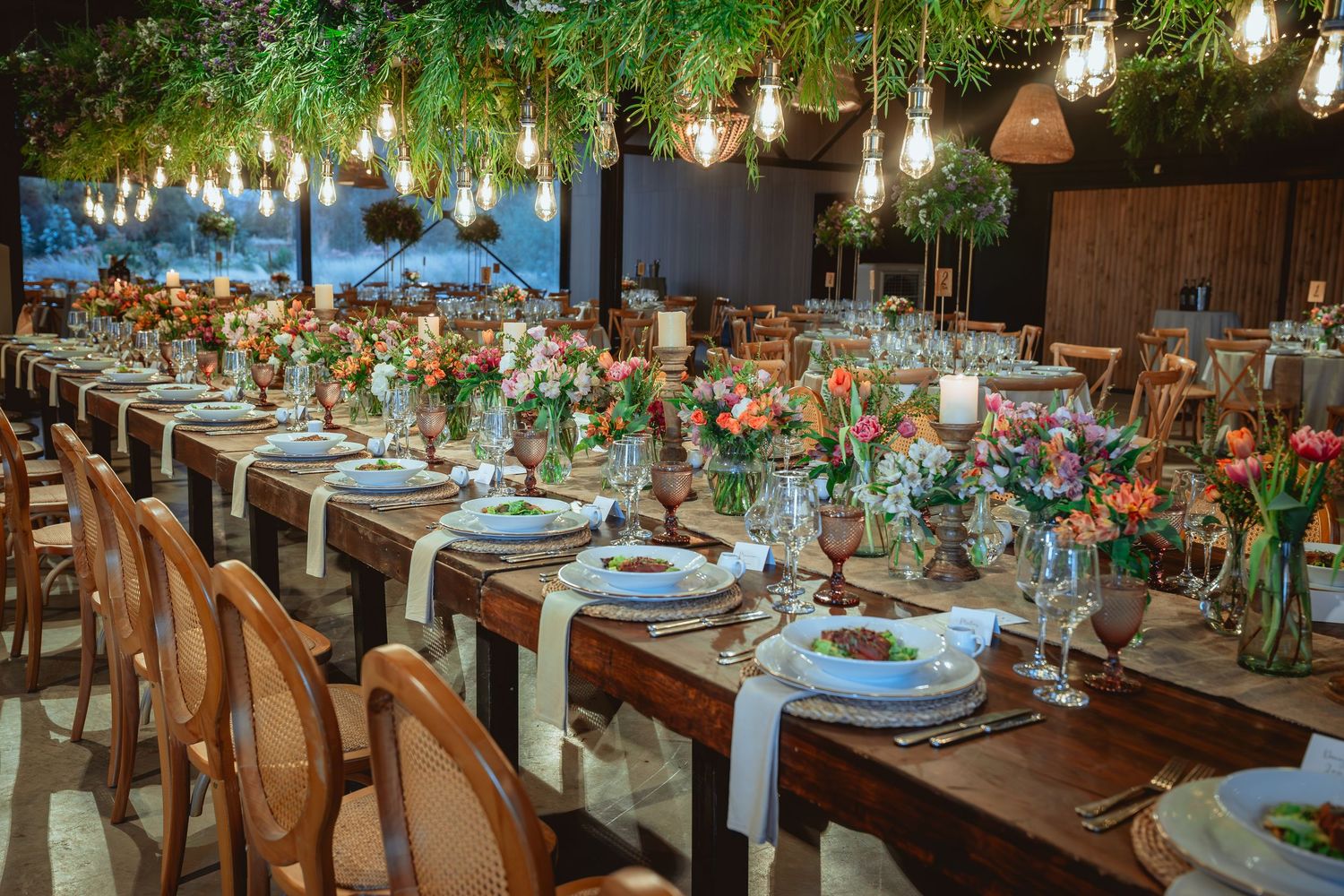 Stunning indoor wedding banquet with long wooden tables, hanging bulb lights, and colorful florals.