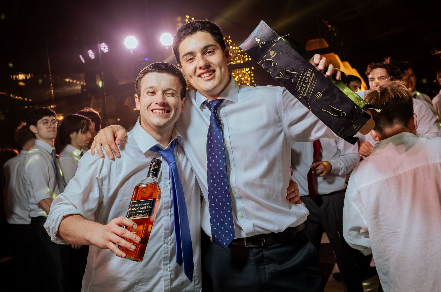Two smiling men in ties hold whiskey bottle at lively wedding reception party.