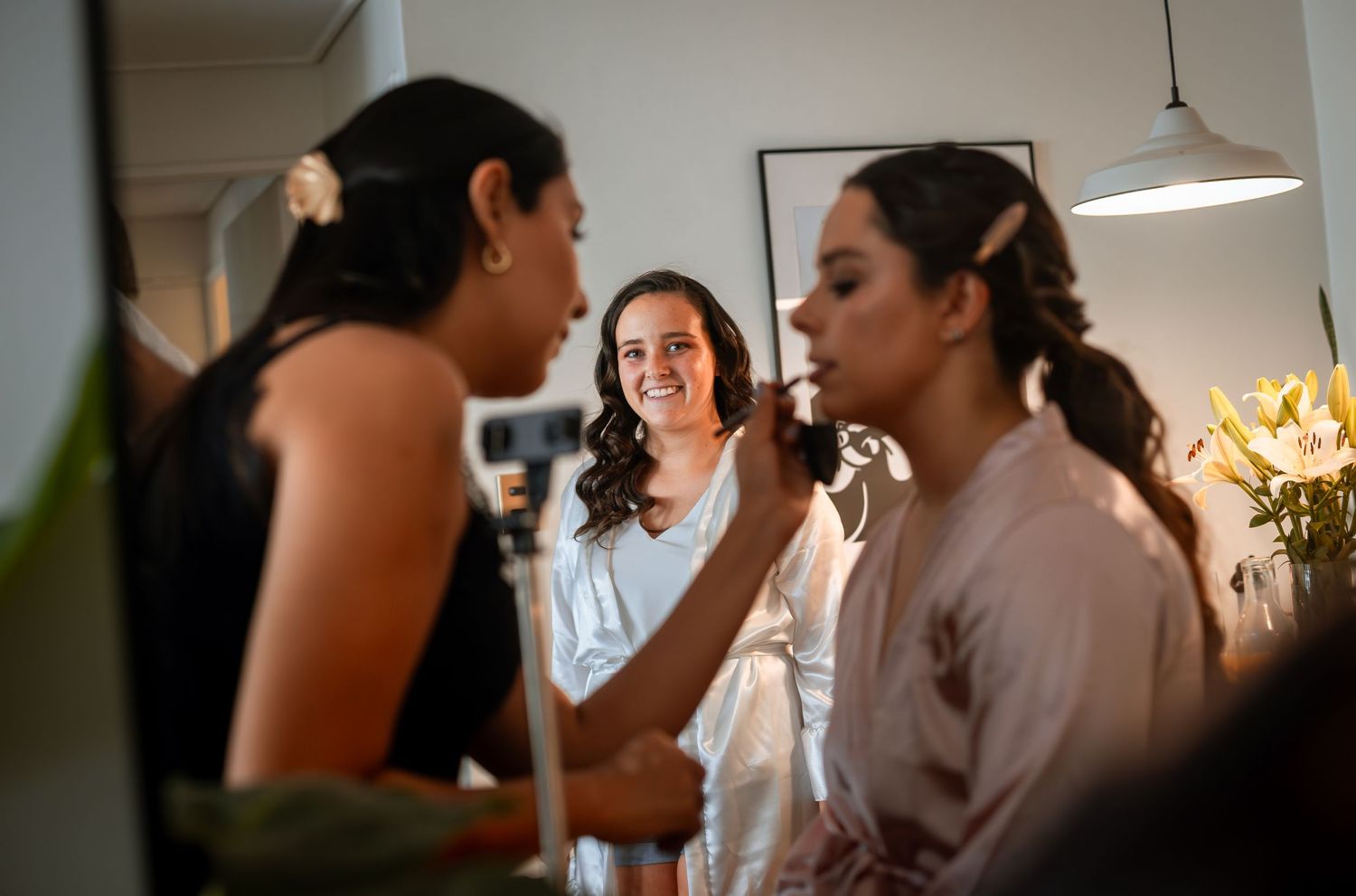 Bride smiling as bridesmaids assist with makeup and hair during wedding day preparations indoors.