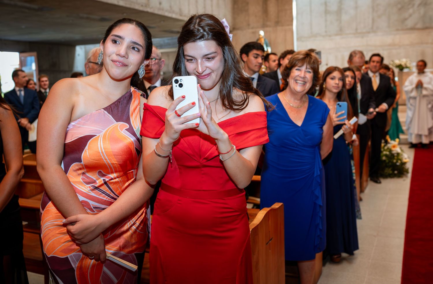 Women in colorful dresses photograph wedding procession on smartphone in church.