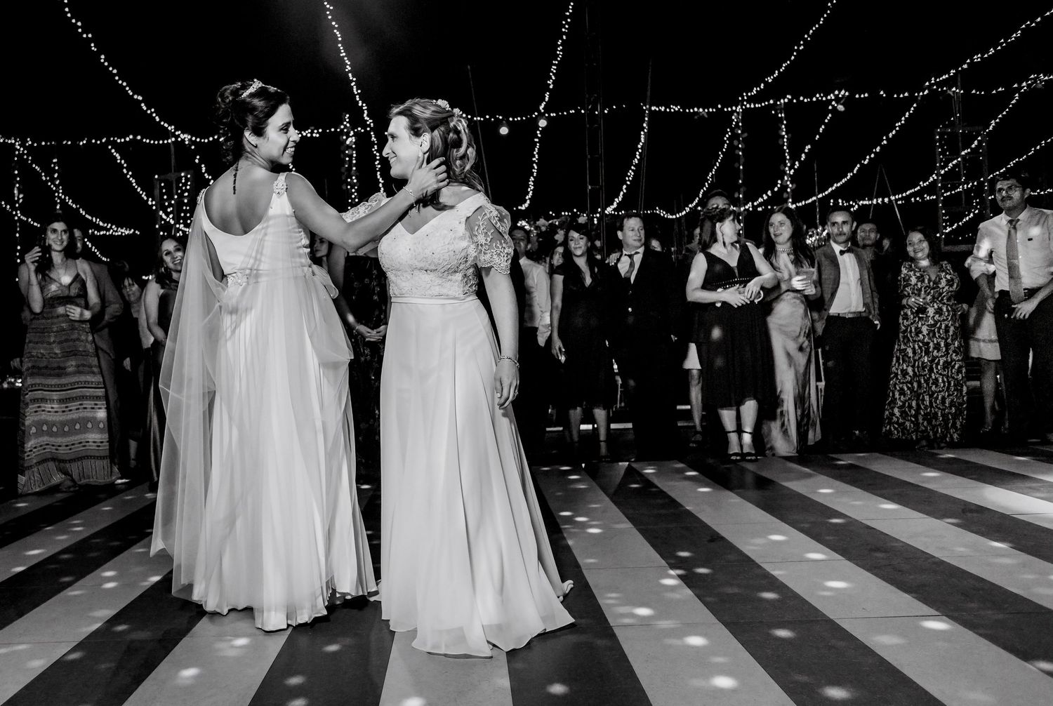 Two brides share first dance on striped floor surrounded by guests in black and white photo.
