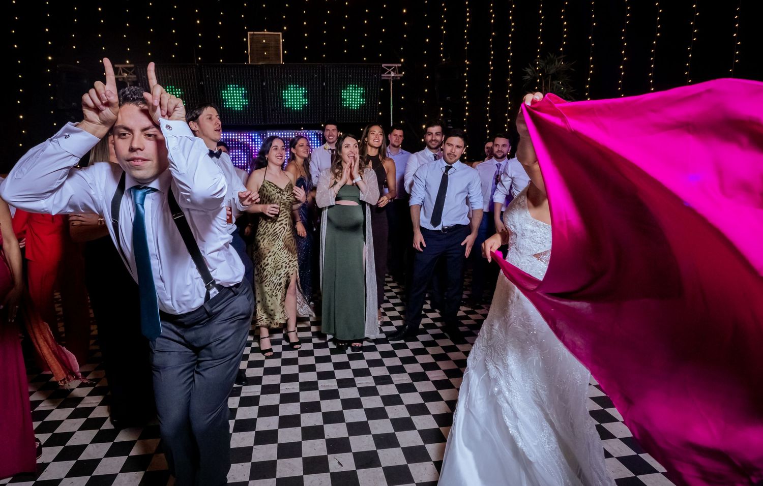 Wedding guests dance and celebrate on a checkered floor with the bride visible in the background.