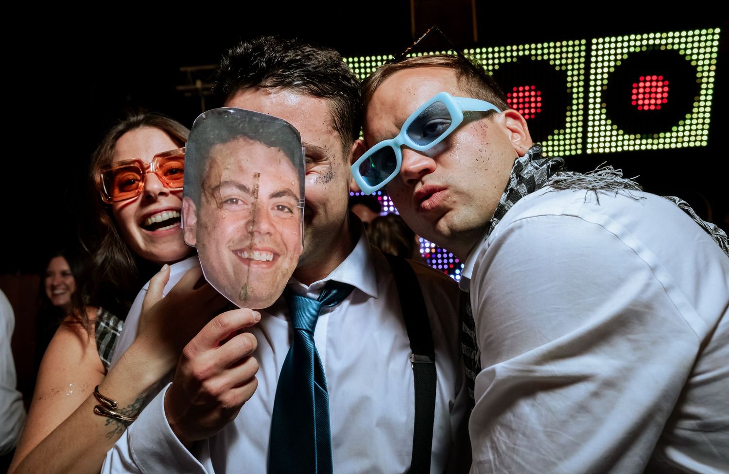 Wedding guests pose with a cardboard face cutout and novelty glasses at a lively reception party.