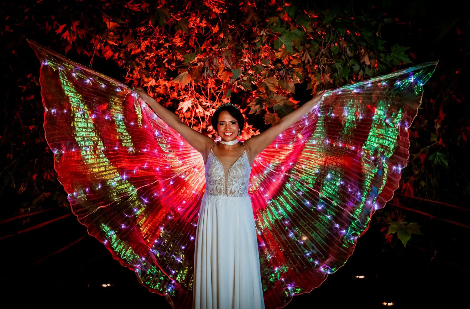 Woman in white dress smiling with large glowing red and green LED butterfly wings outdoors at night.
