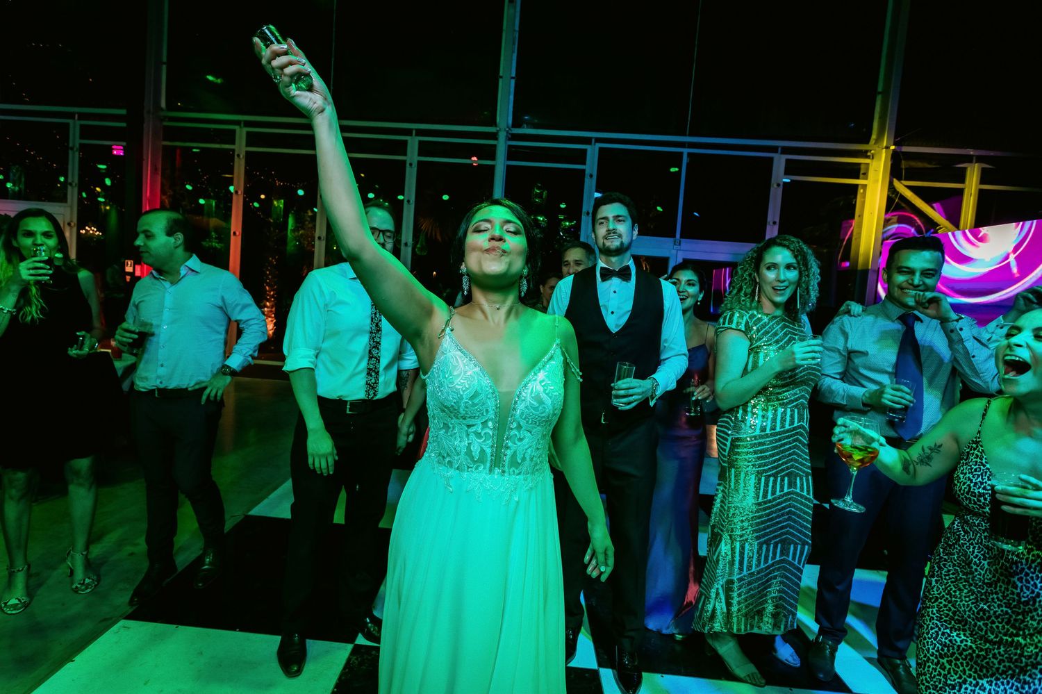 Bride in white dress raises arm celebrating on illuminated dance floor at vibrant wedding reception.