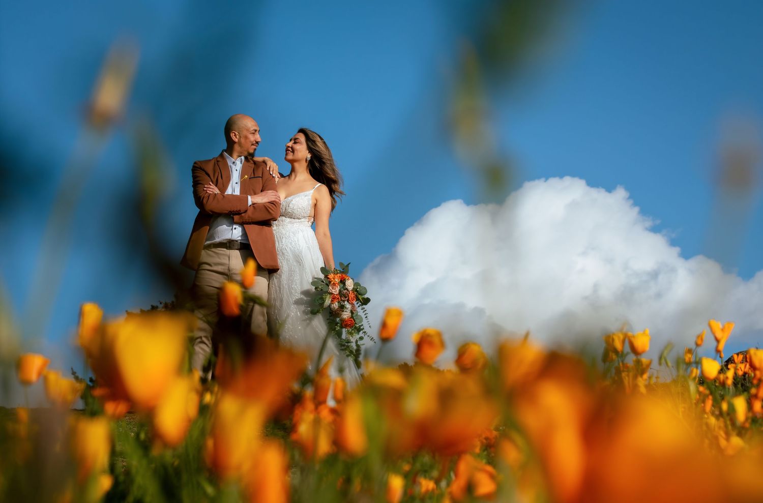Smiling couple embracing among blurred orange wildflowers, the bride holding a bouquet, under a bright blue sky with white clouds.