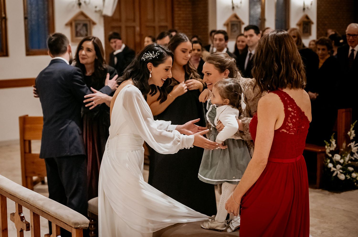 Bride in white gown greets flower girl at church wedding ceremony with guests in background.