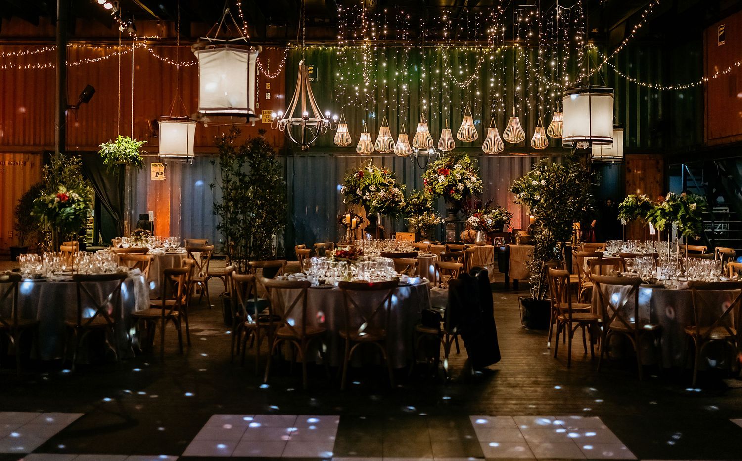 Elegant wedding reception venue with string lights, lanterns, floral arrangements, and a glowing dance floor in a dark, romantic setting.