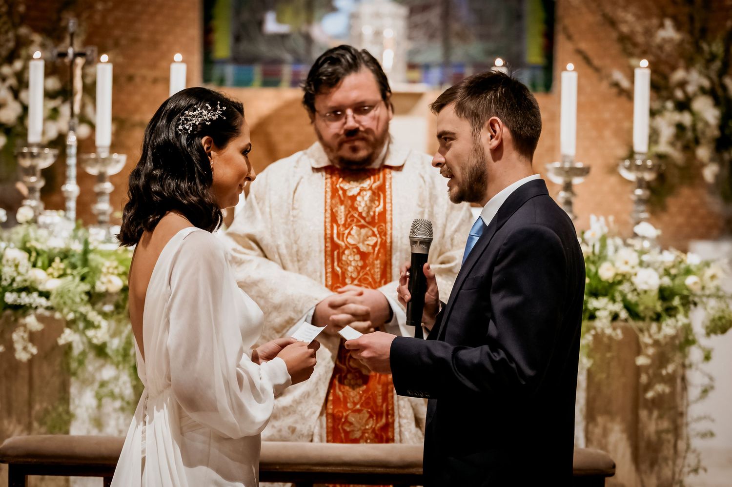 Bride and groom exchange vows before an officiant at a church altar adorned with white flowers and candles.