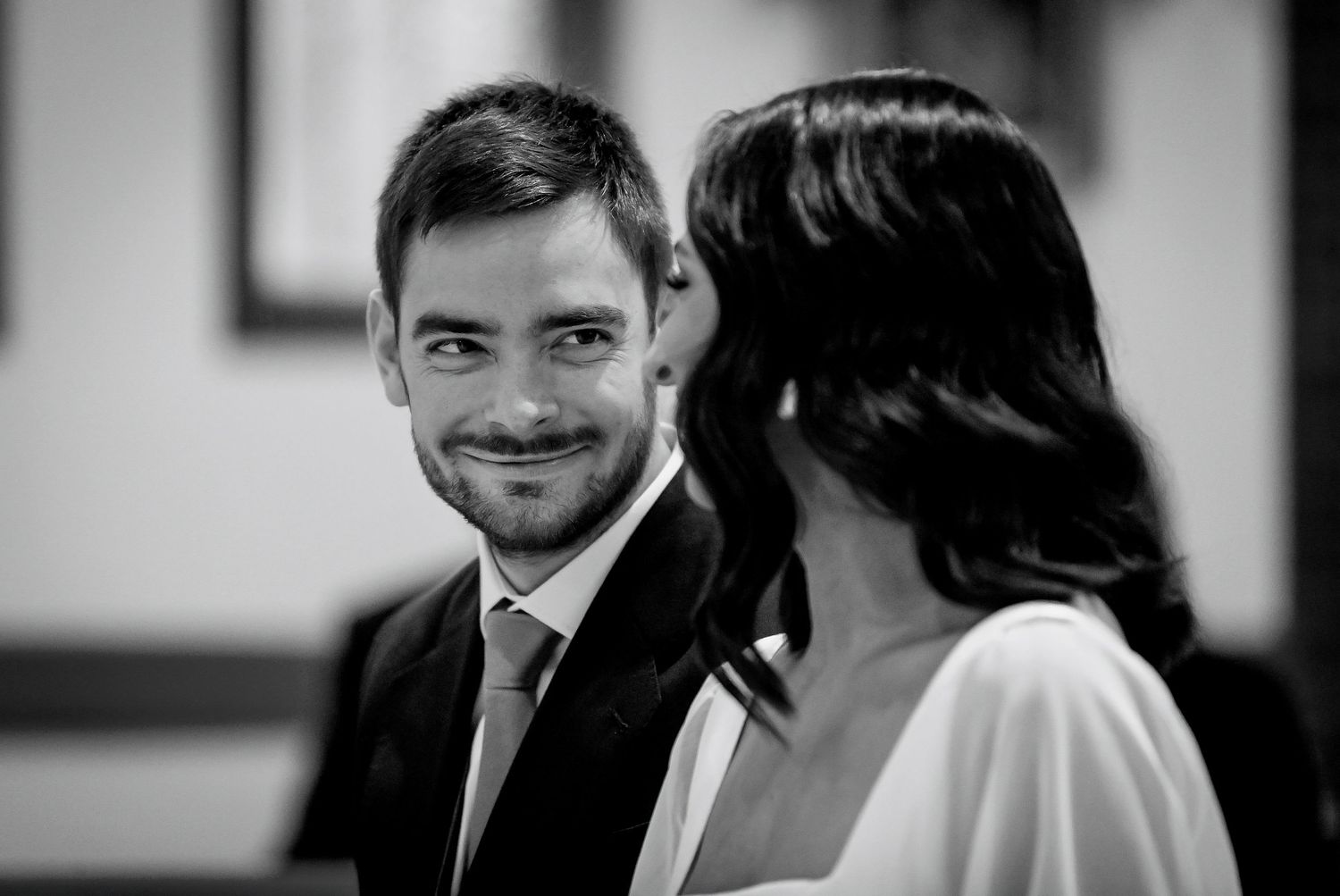 Black and white wedding photo of a smiling groom gazing at his bride during their ceremony.