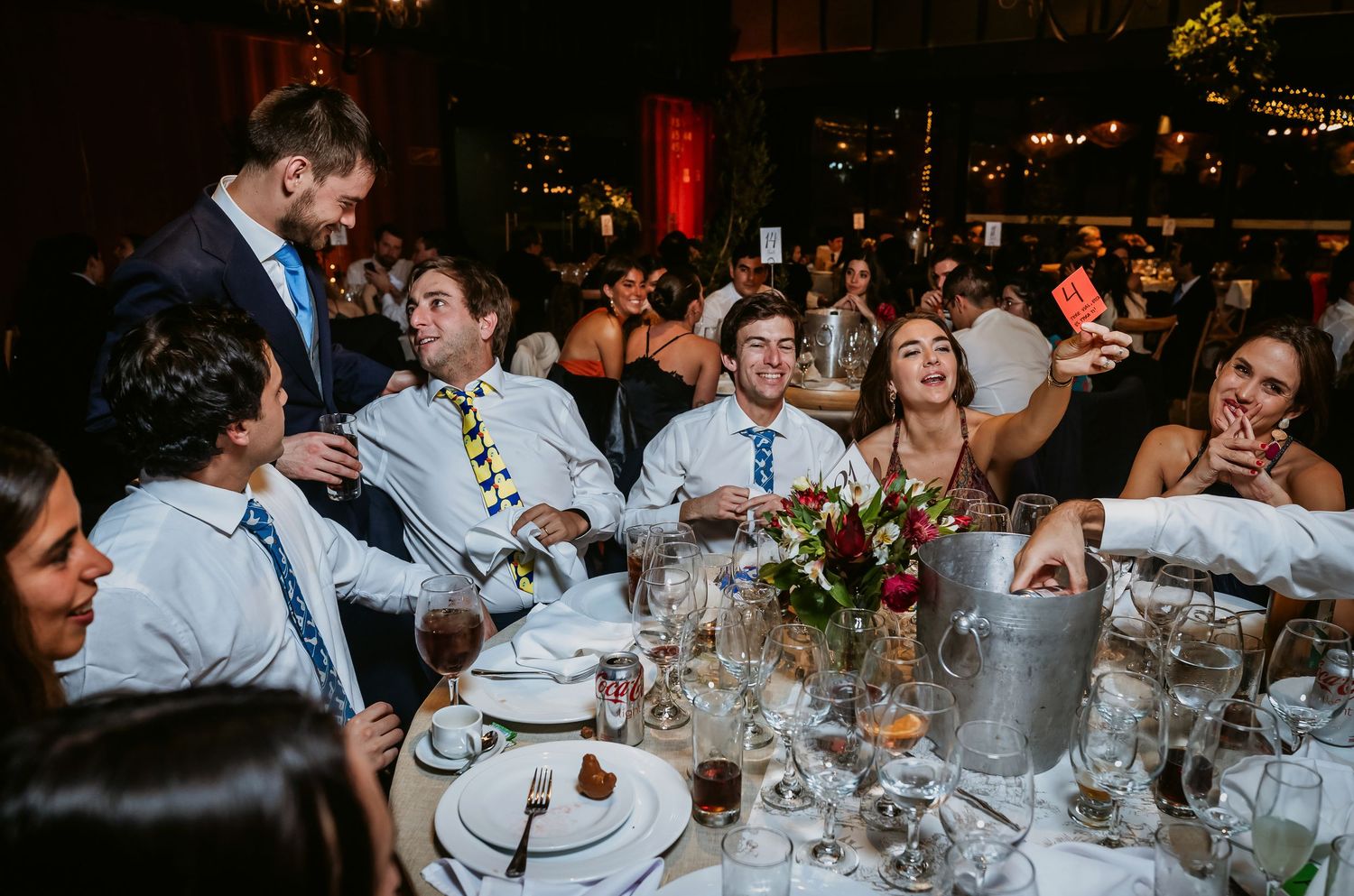 Guests laughing and socializing at a festive wedding reception dinner table with floral centerpieces and dim lighting.