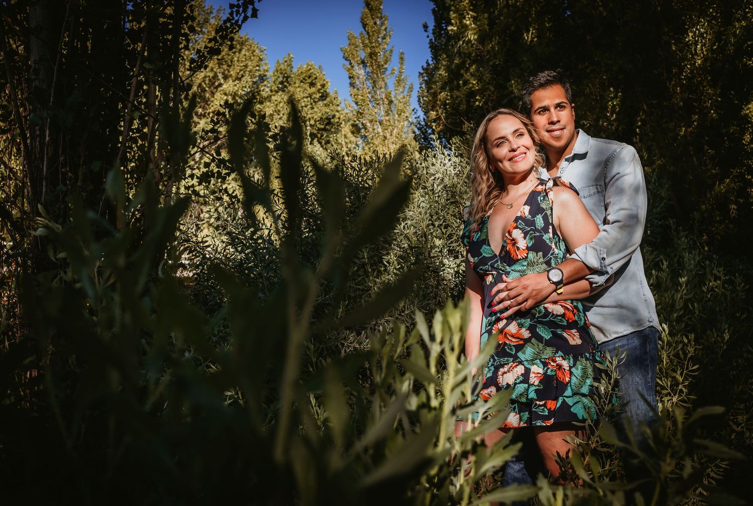 Couple embracing romantically among lush green plants and trees in an outdoor garden setting.
