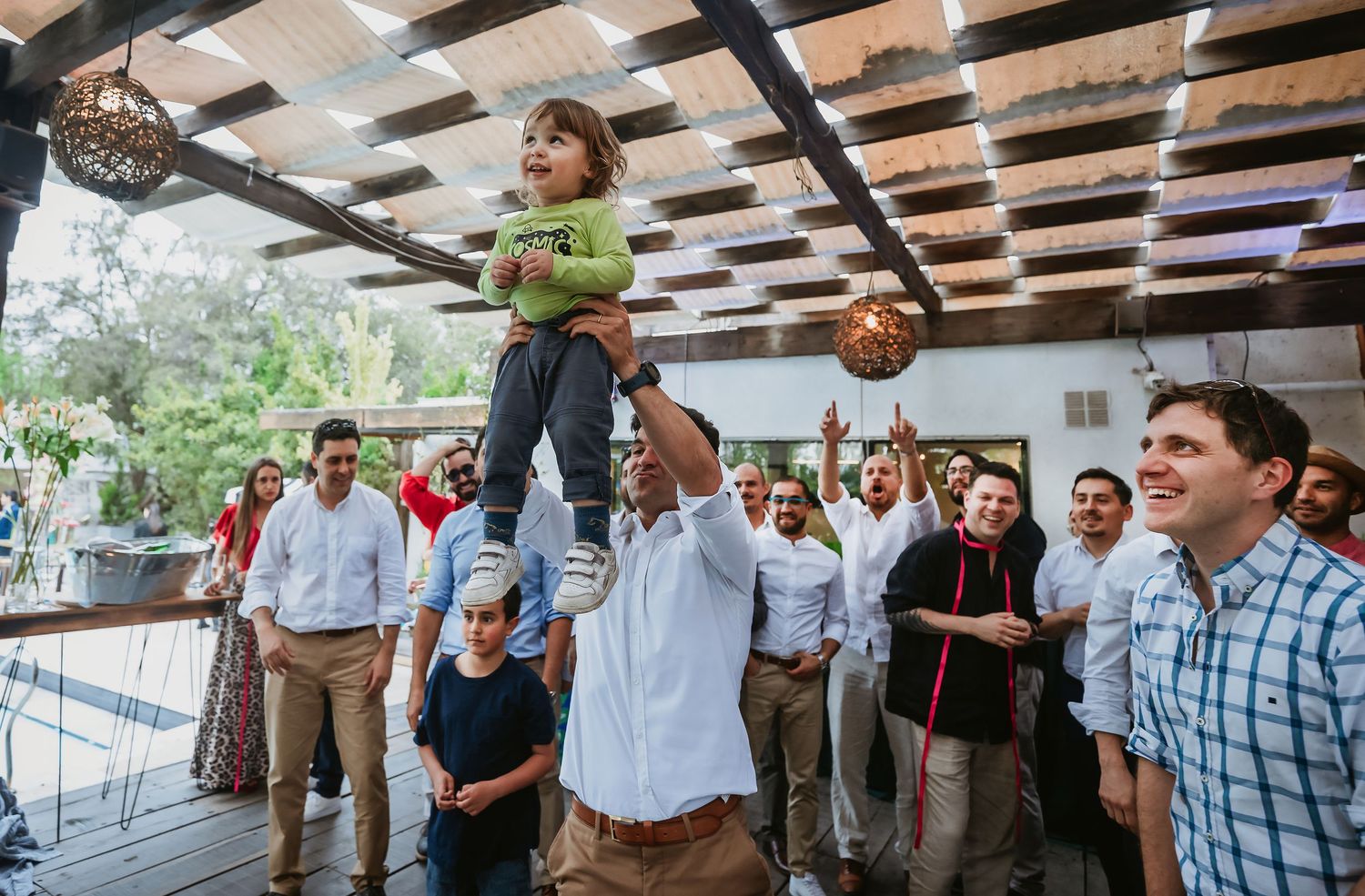 Man lifting a joyful child above a crowd at an outdoor celebration with guests cheering in the background.
