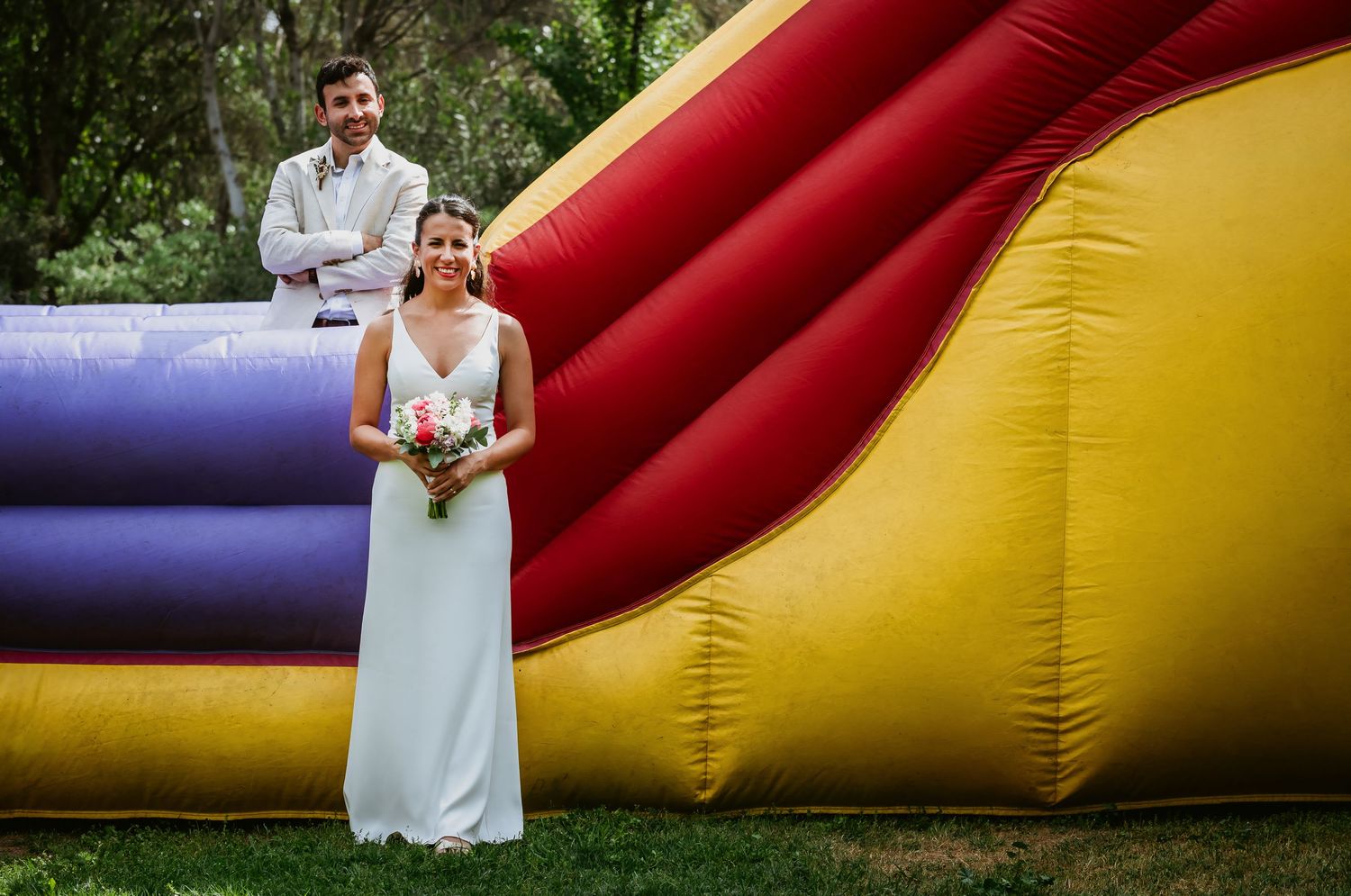 Bride in white gown holds bouquet beside colorful inflatable bounce house as groom stands on top smiling.