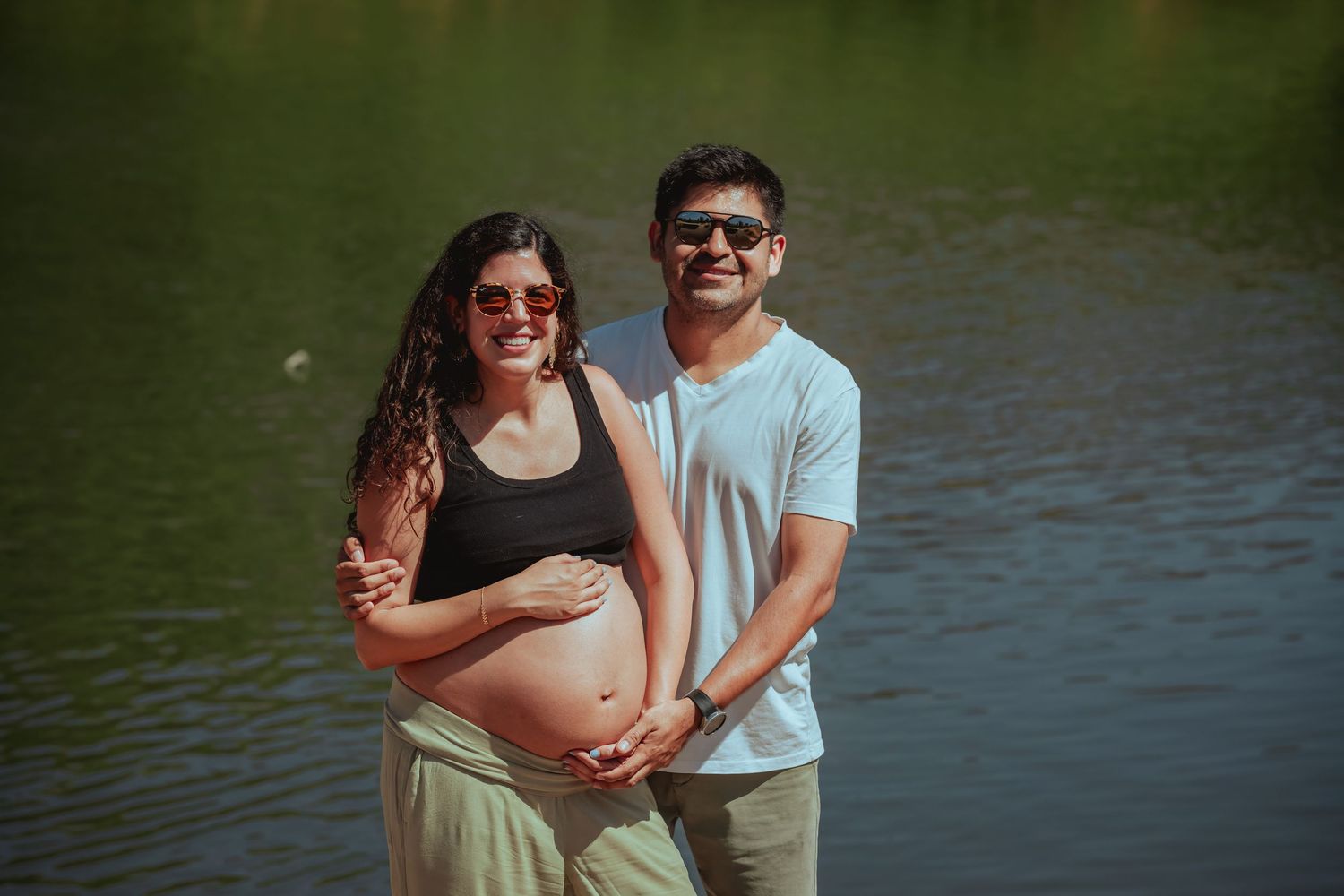 Pregnant couple smiling by a lake, woman in black crop top cradling baby bump, man in blue shirt wearing sunglasses.